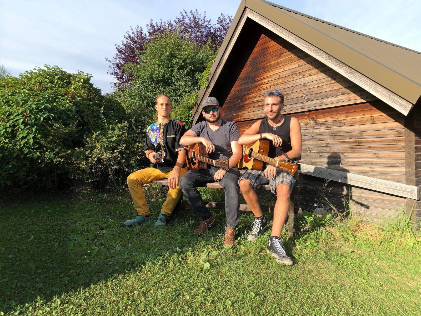 Simon, Max und Markus auf einer Bank vor einer Holzhütte im Abendlicht, zwei von ihnen mit Gitarre